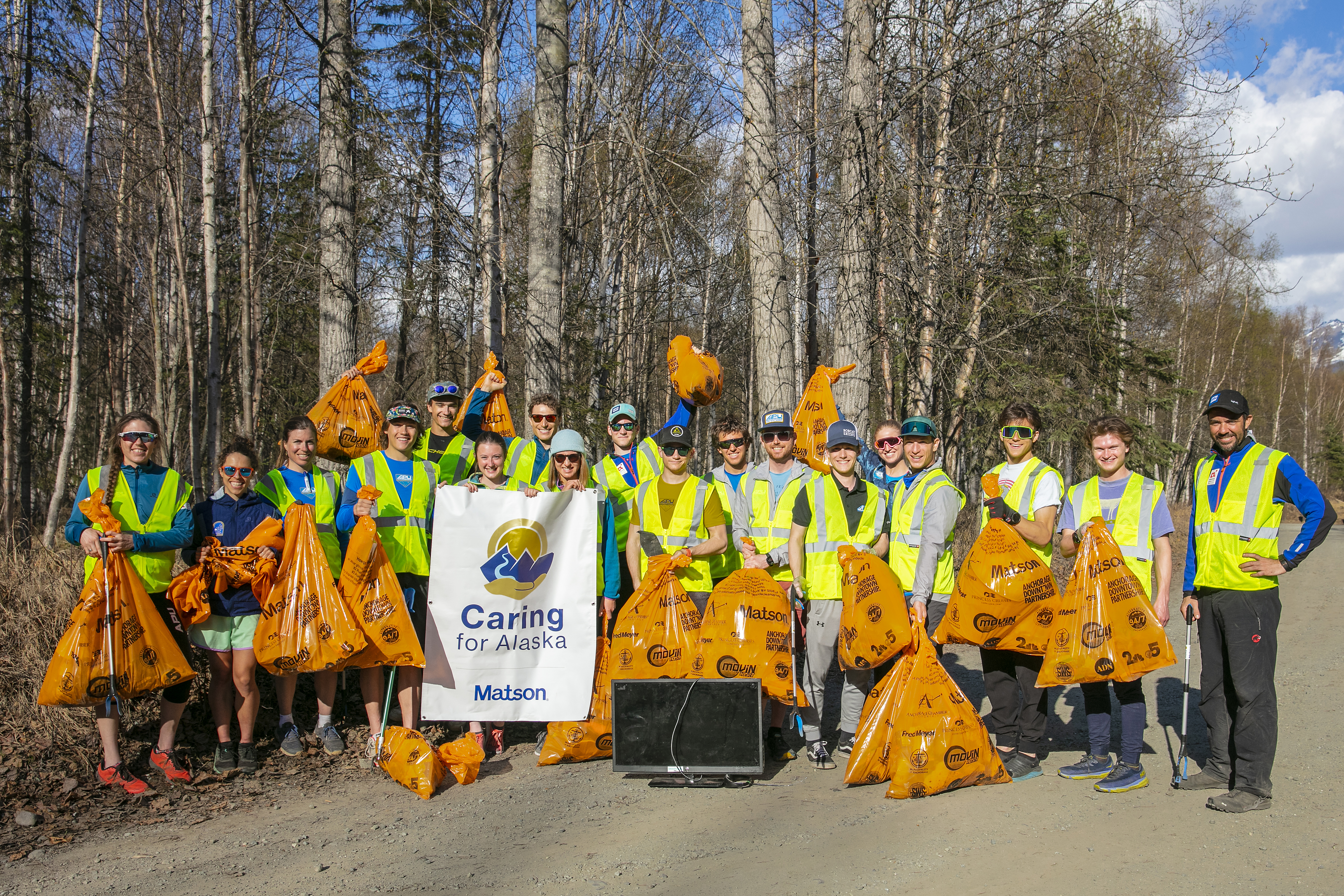Volunteers wearing yellow safety vests pose for a group picture with their loaded orange trash bags and Caring For Alaska banner.