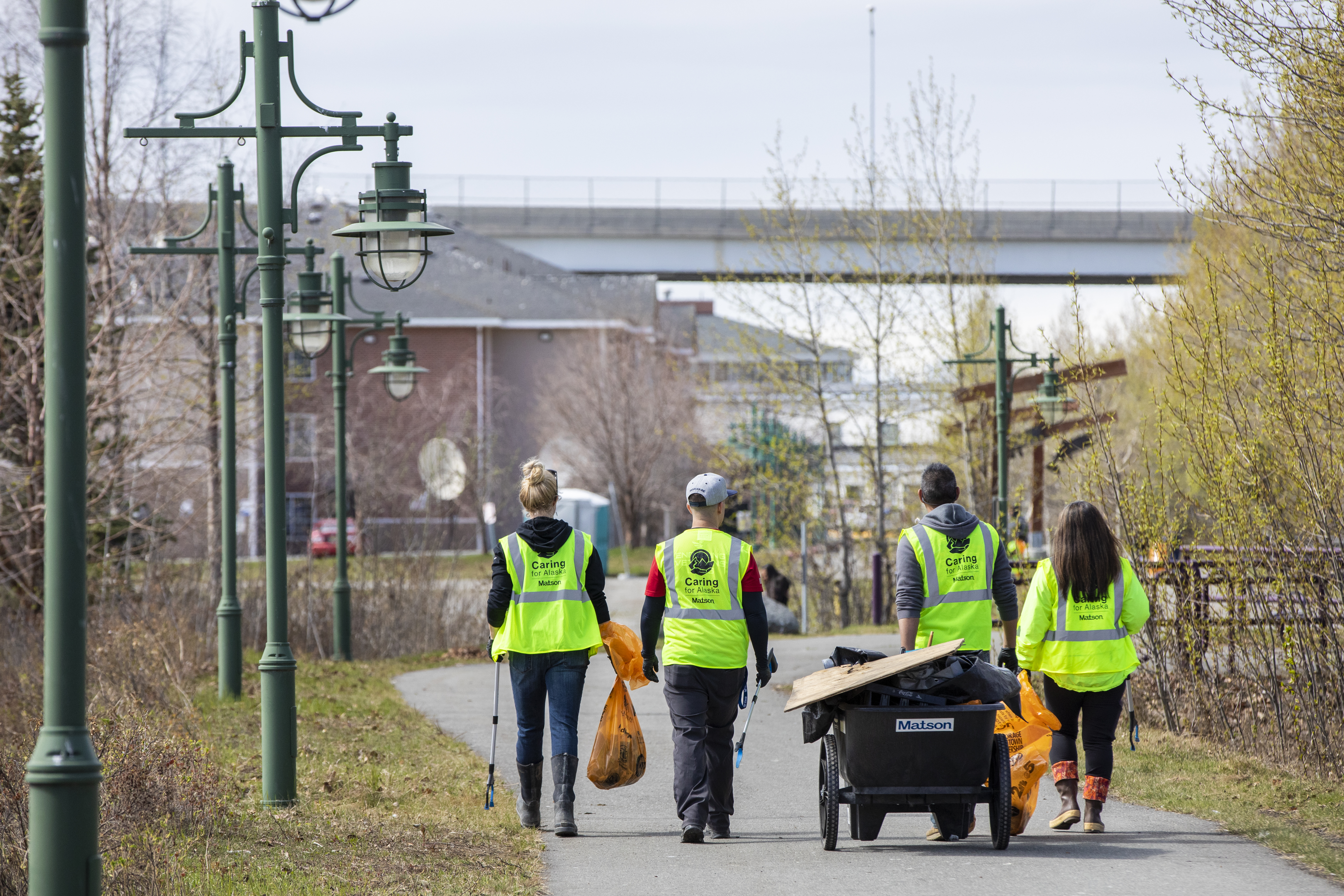 Volunteers wearing yellow safety vests walk along Ship Creek with a cart and orange bags full of trash.