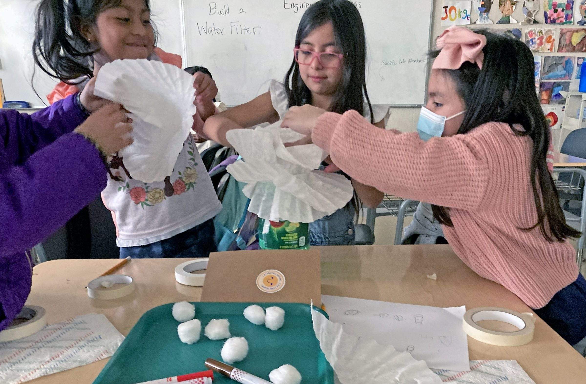 Girls stack paper coffee filters on a can in a classroom. Masking tape, cotton balls, paper towels, and markers sit on their table.