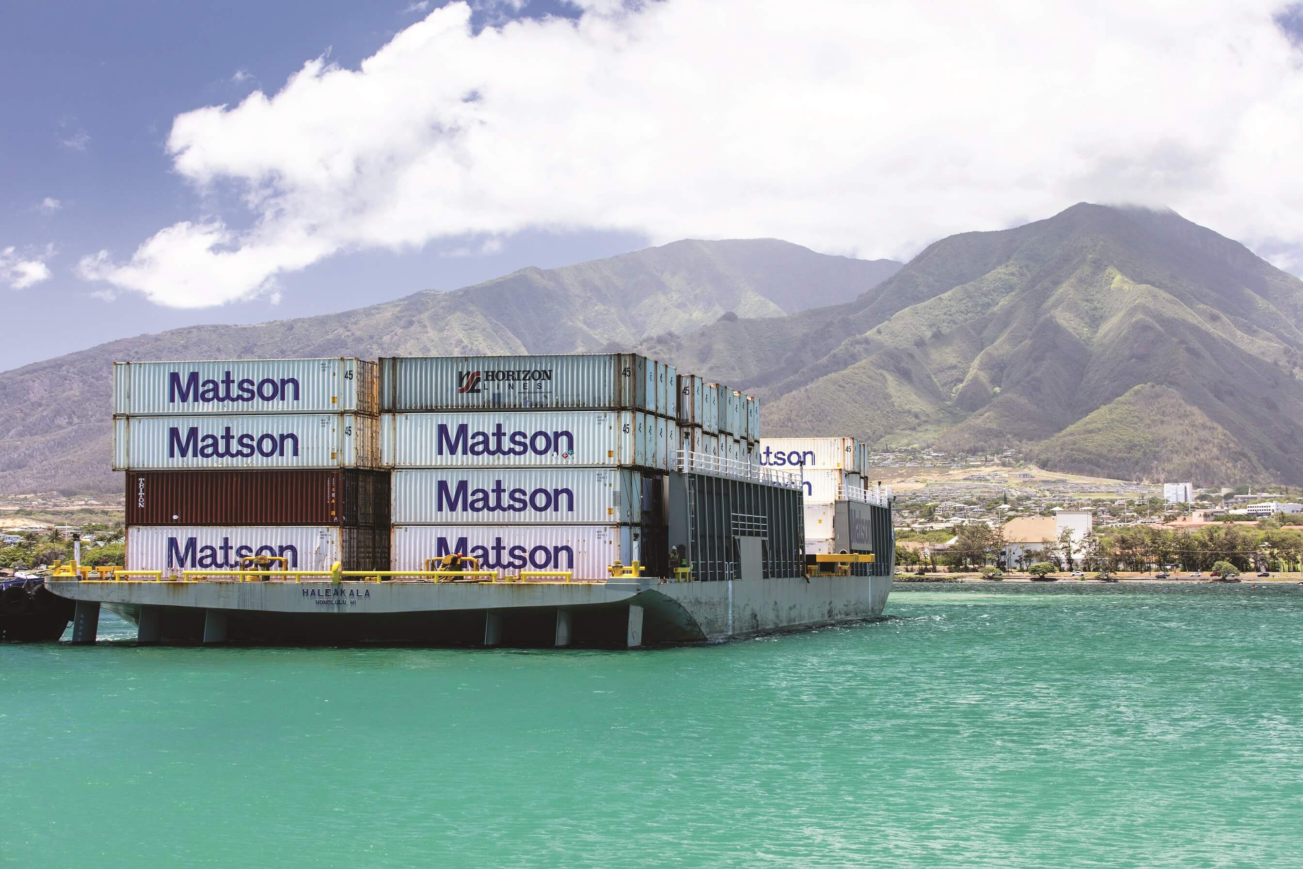 Matson barge Haleakala loaded with Matson containers arrives in Kahului with green mountains and clouds in the background.