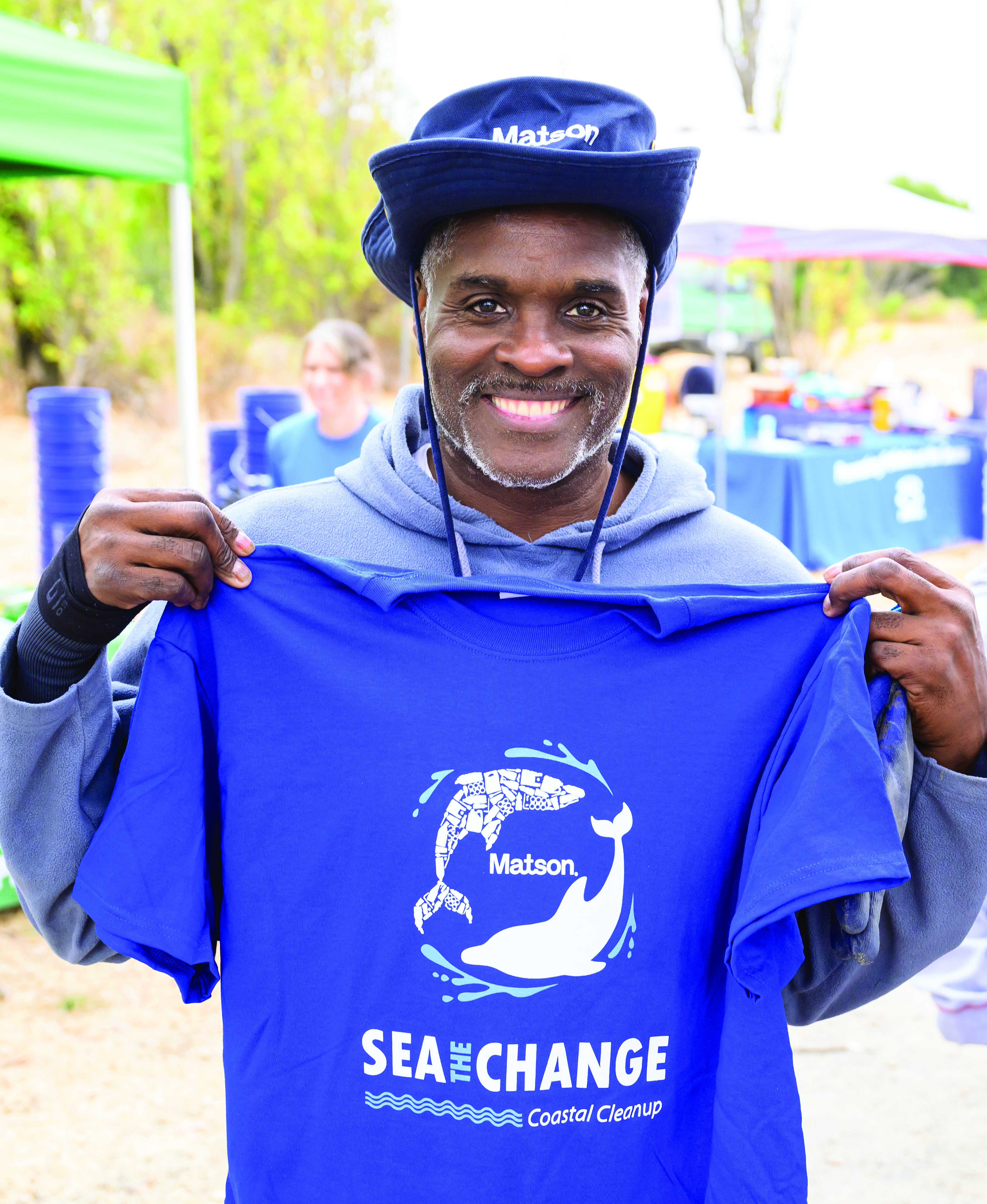 Volunteer wears a blue Matson branded hand and holds up a blue t-shirt printed with two dolphins encircling a Matson logo and the words "Sea The Change."