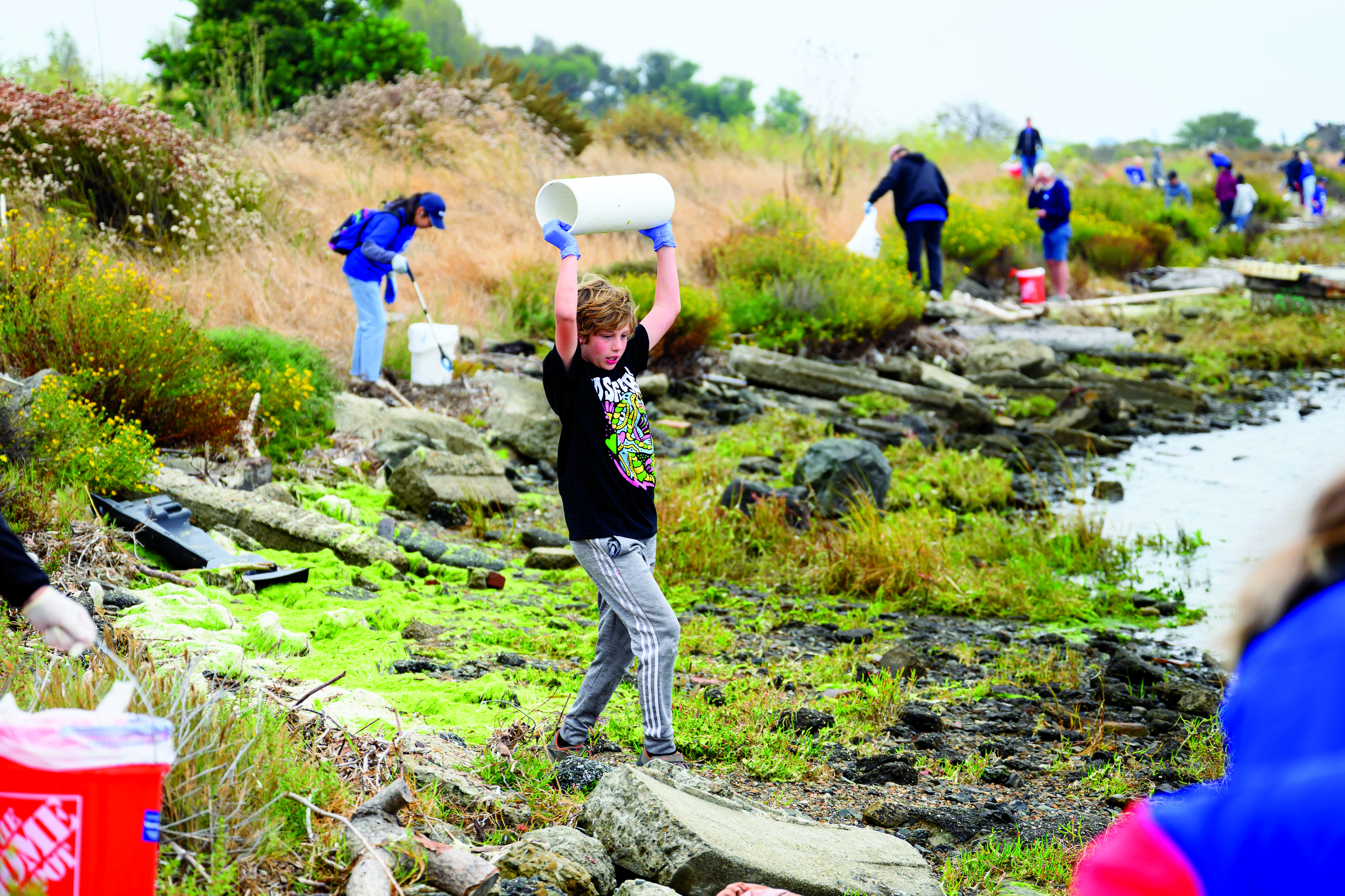 Young male tween wearing blue plastic gloves holds up a large section of plastic piping at the estuary wtih other clean up volunteers in the background.