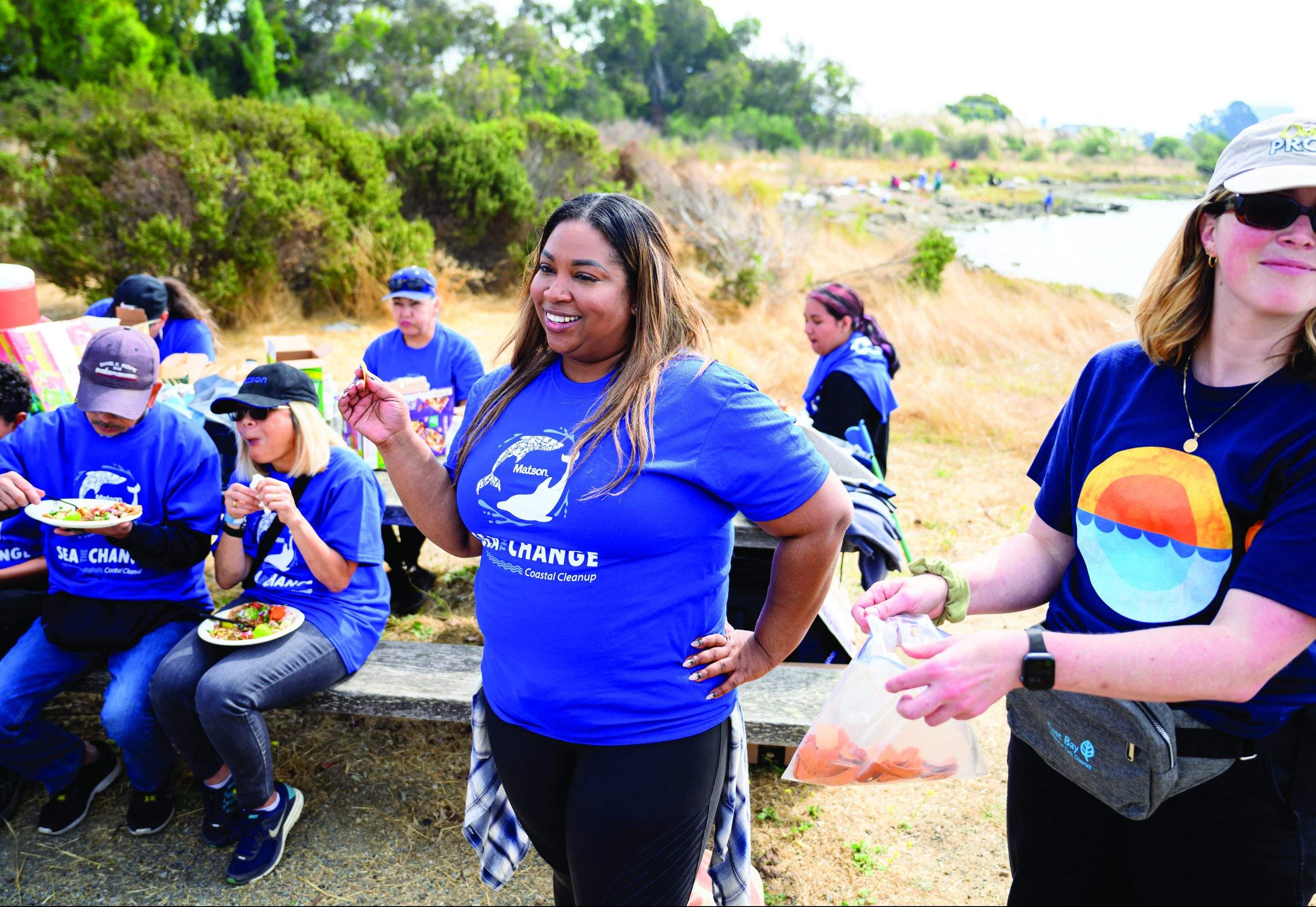 Denise stands with a raffle ticket in hands as volunteers wearing blue Matson "Sea The Change" t-shirts sit on a picnic table behind her eating.