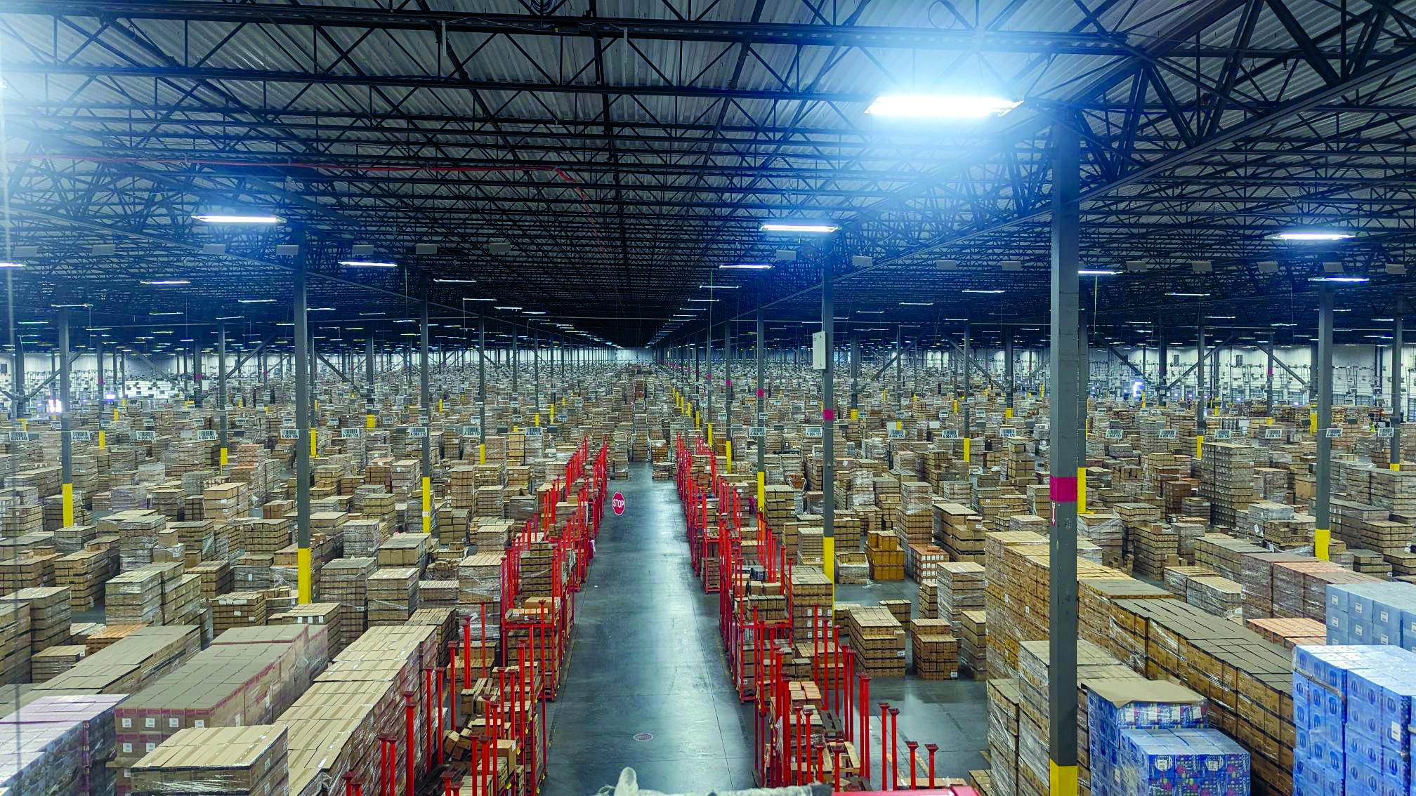 Pallets of brown boxes sit stacked in an expansive high ceiling warehouse.