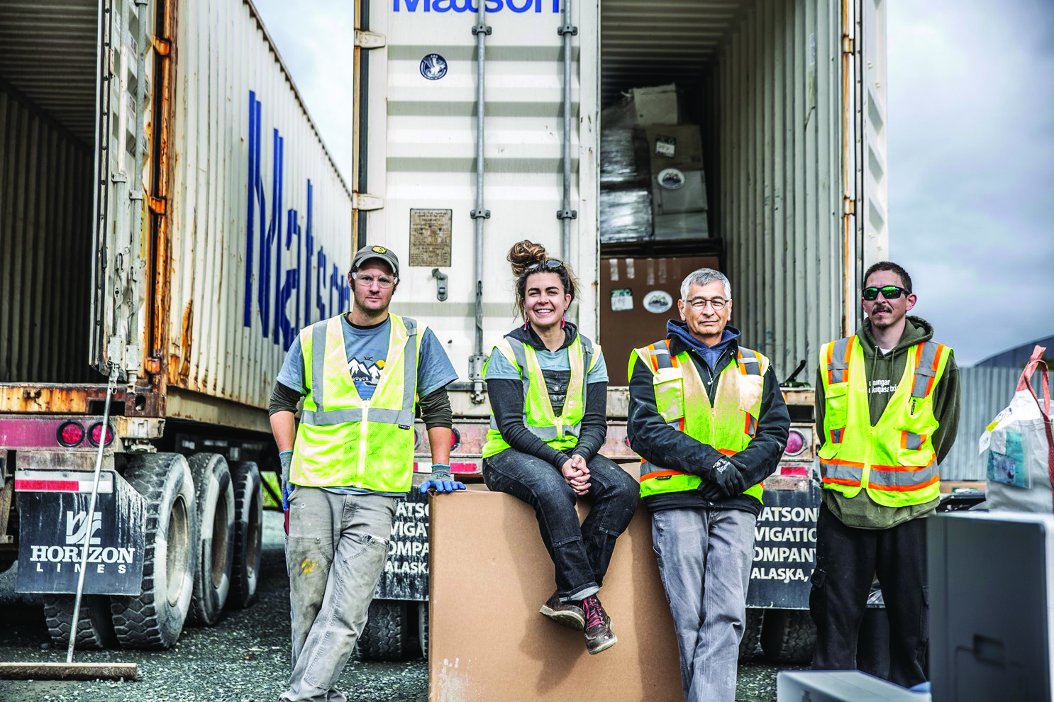 Four Backhaul stafff and volunteers wearing yellow safety vests pose in front of a Matson container loaded with e-waste.