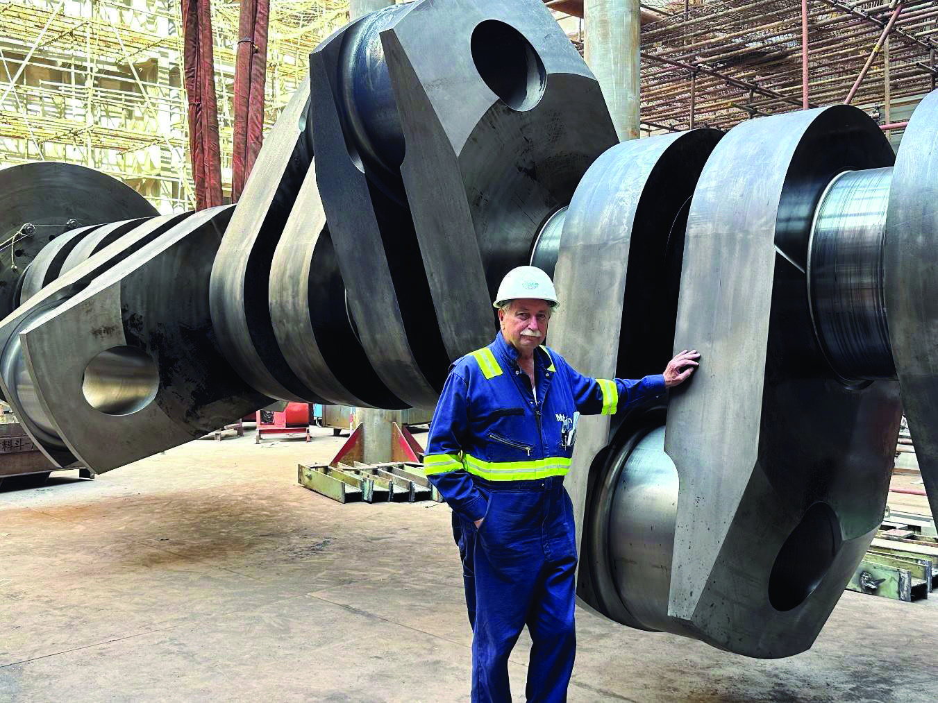 Angus Lundstrum, wearing a white hardhat and blue jumpsuit with yellow safety stripes, is dwarfed next to the Manukai's crankshaft.
