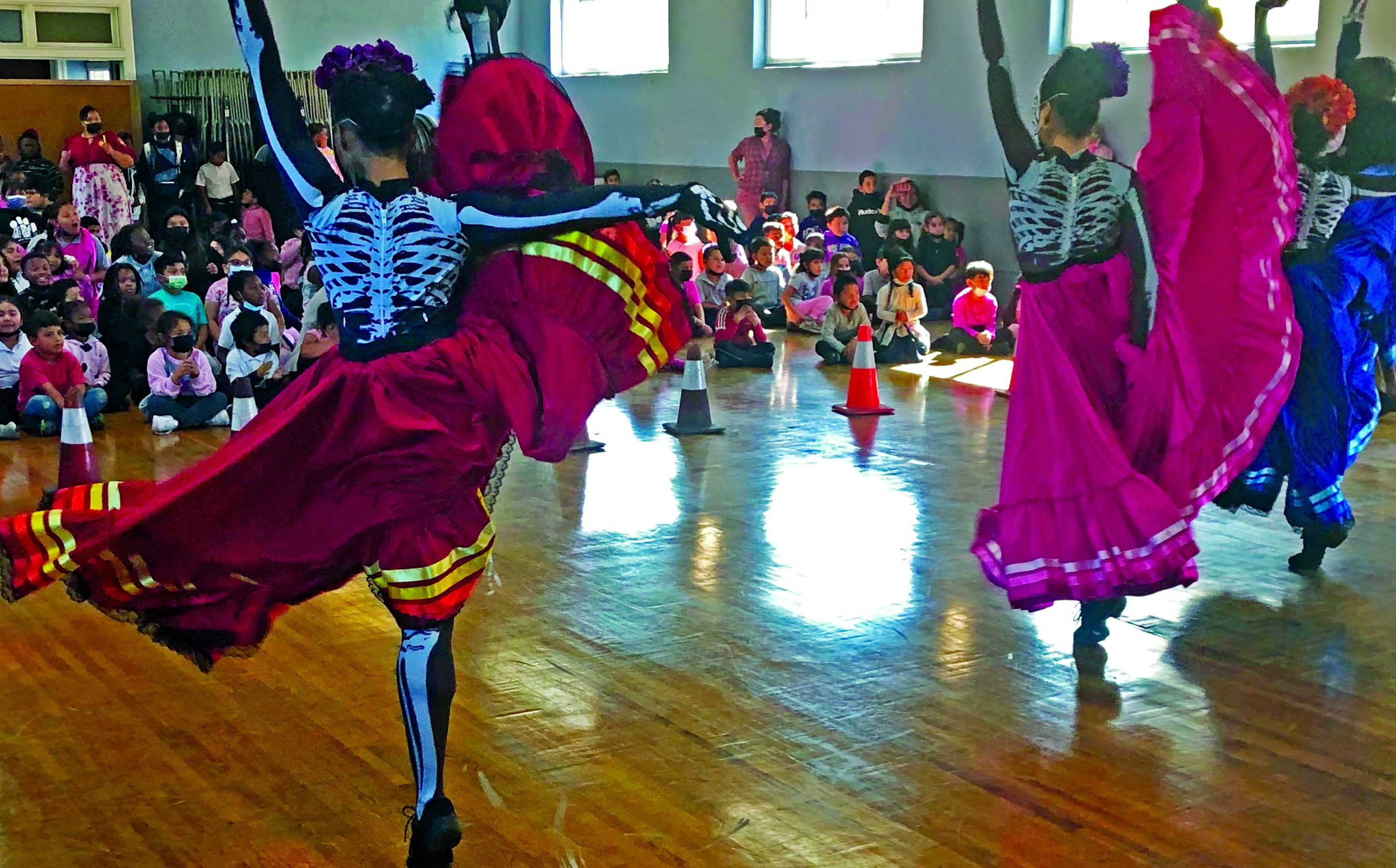 Female dancers, wearing leotards deocrated with a skeleton and colorful skirts, kick their right let in the air.