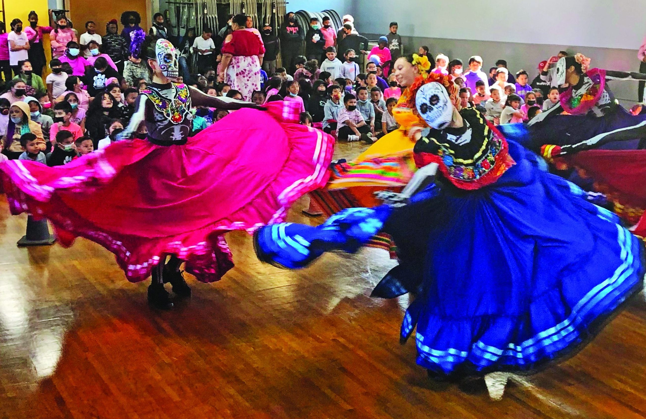 Female dancers, wearing masks decorated as skulls, turn about the room in colorful, full skirts as students watch.