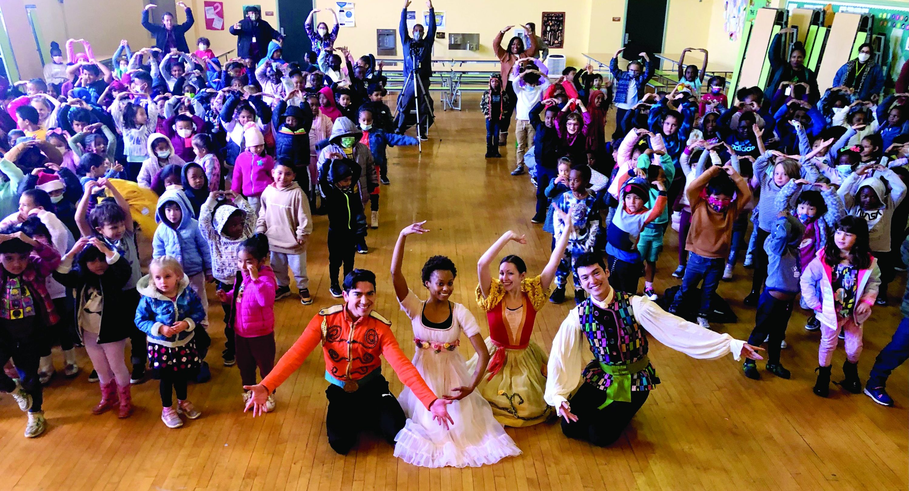 Four performers from OBC's "The Nutcracker" pose in the middle of the aisle while elementary students hold their arms above their head with fingers touching.
