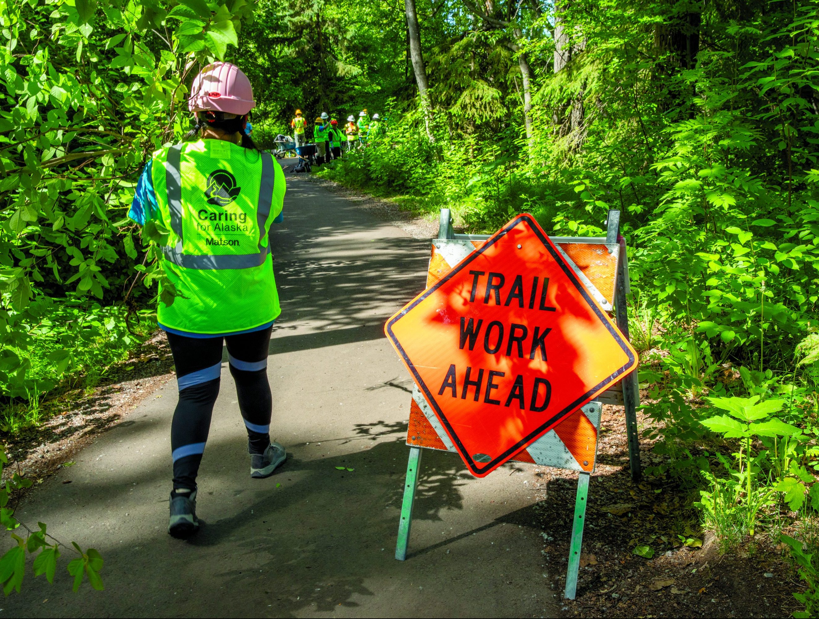 An Anchorage Park Foundation’s Youth Employment in Parks participant wearing a yellow Caring for Alaska vest walks past a Trail Work Ahead sign on a paved park path.