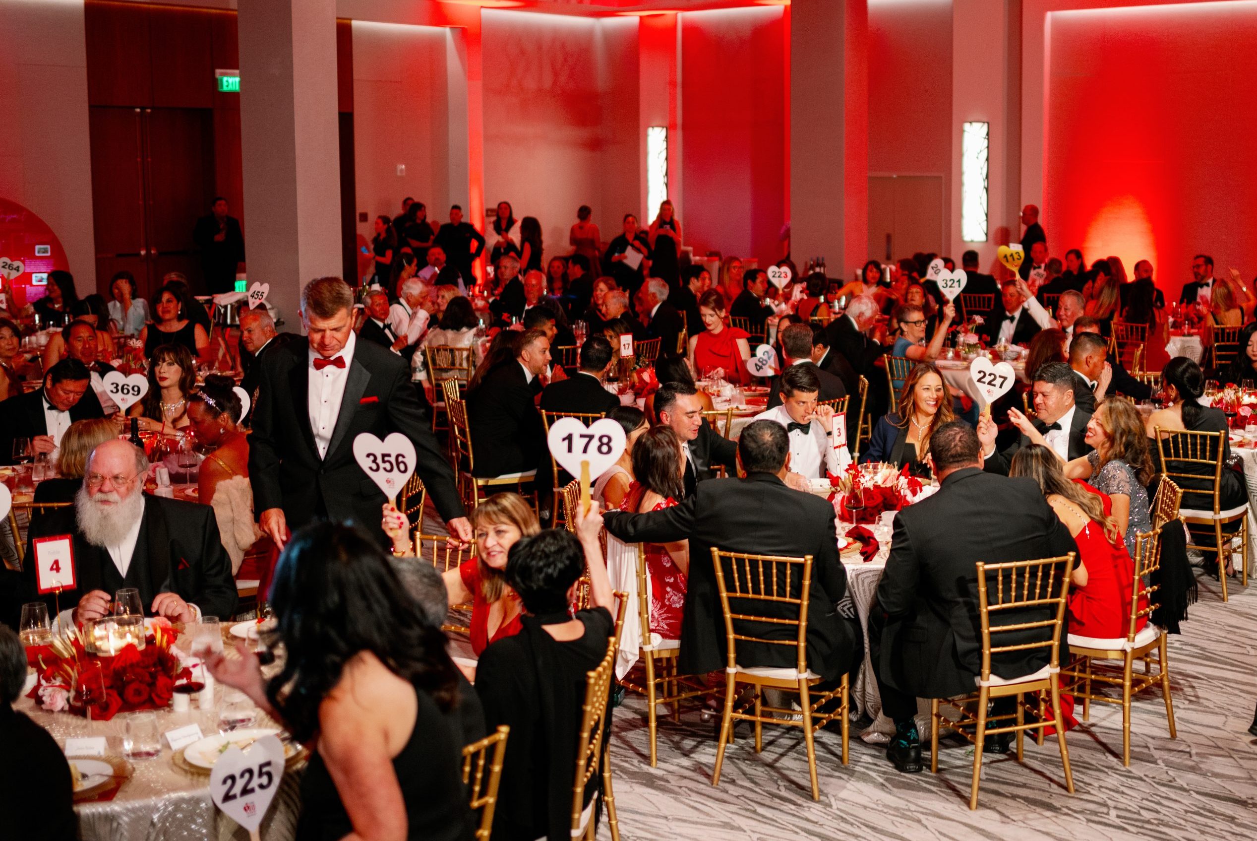 Heart Ball suppoters wearing tuxedos and dresses sit at round tables with heart shaped auction paddles in a hotel ballroom bathed in red light.