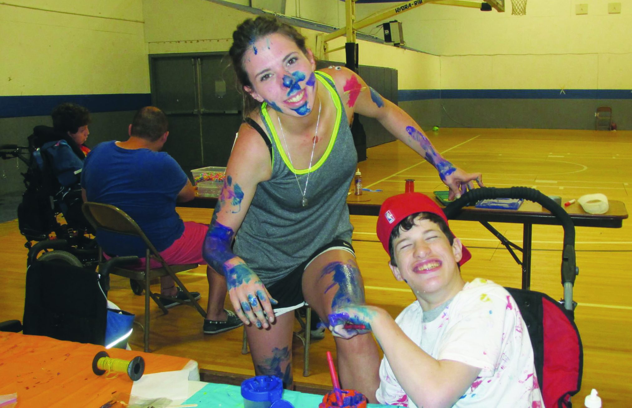 A happy camper wearing a red working with paint sits at a table while supervised by a counselor covered in paint from head to legs.