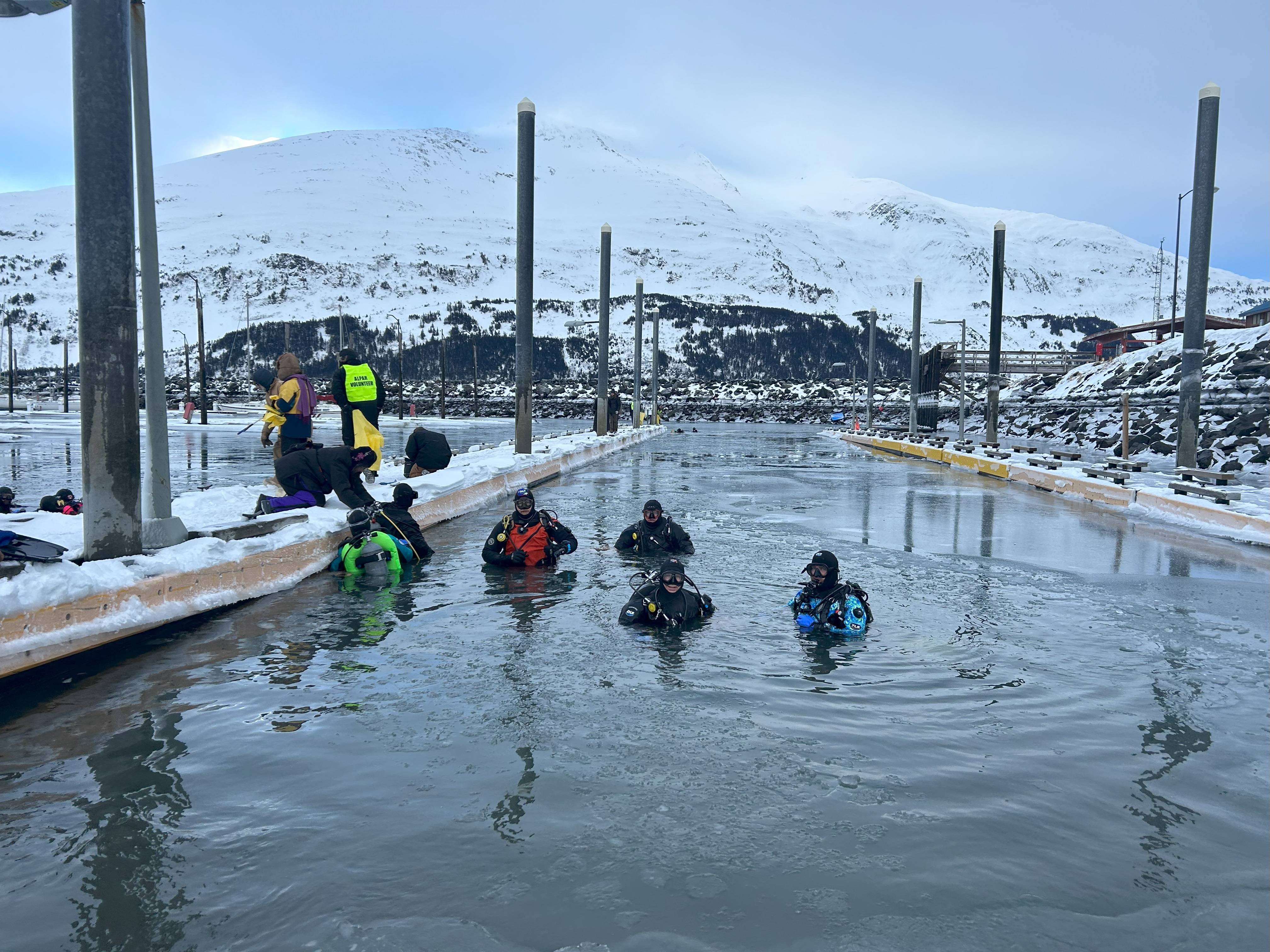 Divers stand in frigid Whittier Harbor waters with a snow capped mountain in the background.