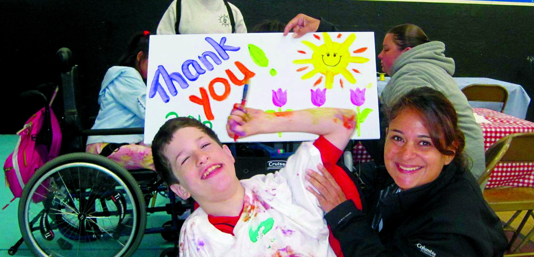 A happy camper wearing a white t-shirt splattered with pain holds a paint brush, while a counselor holds up a painted "thank you" poster with the sun and flowers.