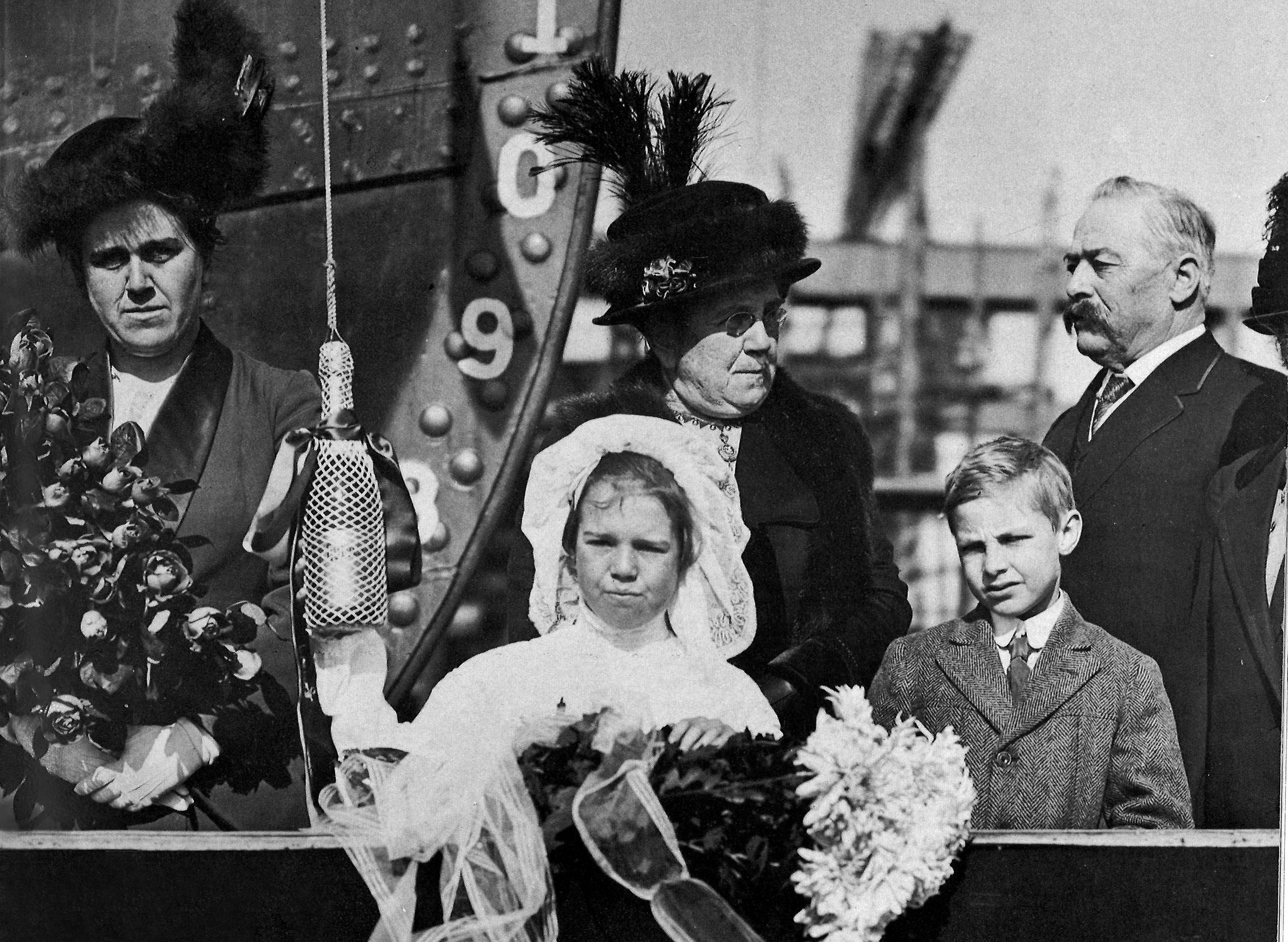 A young girl dressed in white stands ready to break a bottle of champagne hanging in a netted sack as dressed up onlookers wait for the moment.