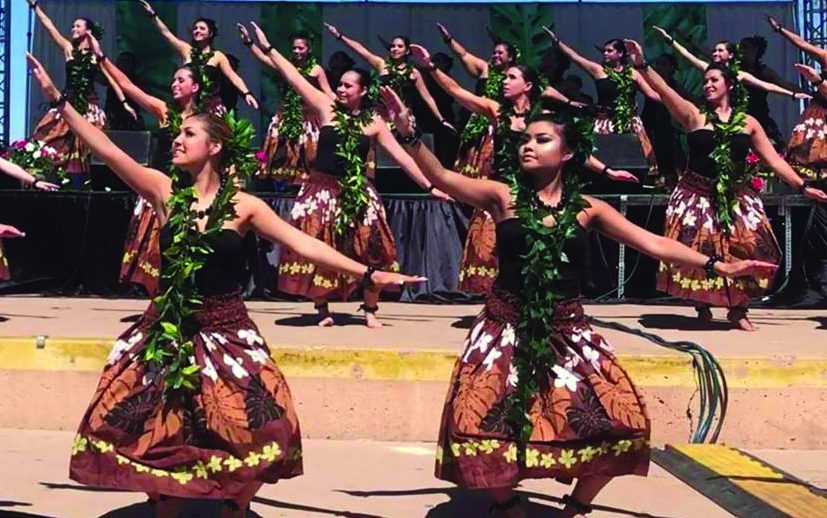 Female dancers adorned with ti leaf leis and brown costumes dance on stage.