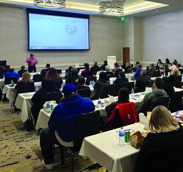 Attendees, facing a screen and speaker, sit in a classroom style arrangement in a conference room.
