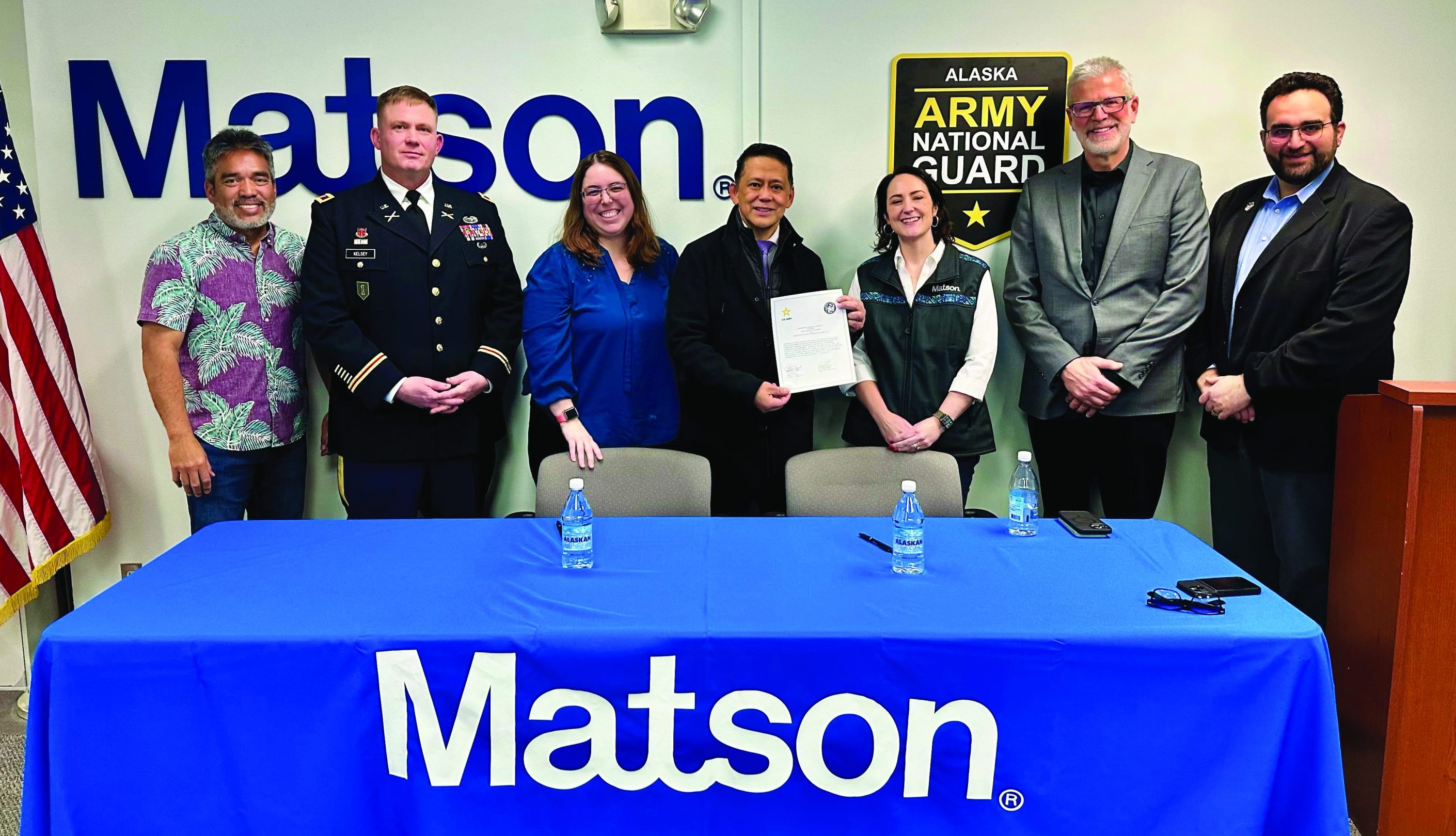 Partners stand behind a table draped with the Matson logo and in front of a wall that has the Matson and Alaska Army National Guard logos.