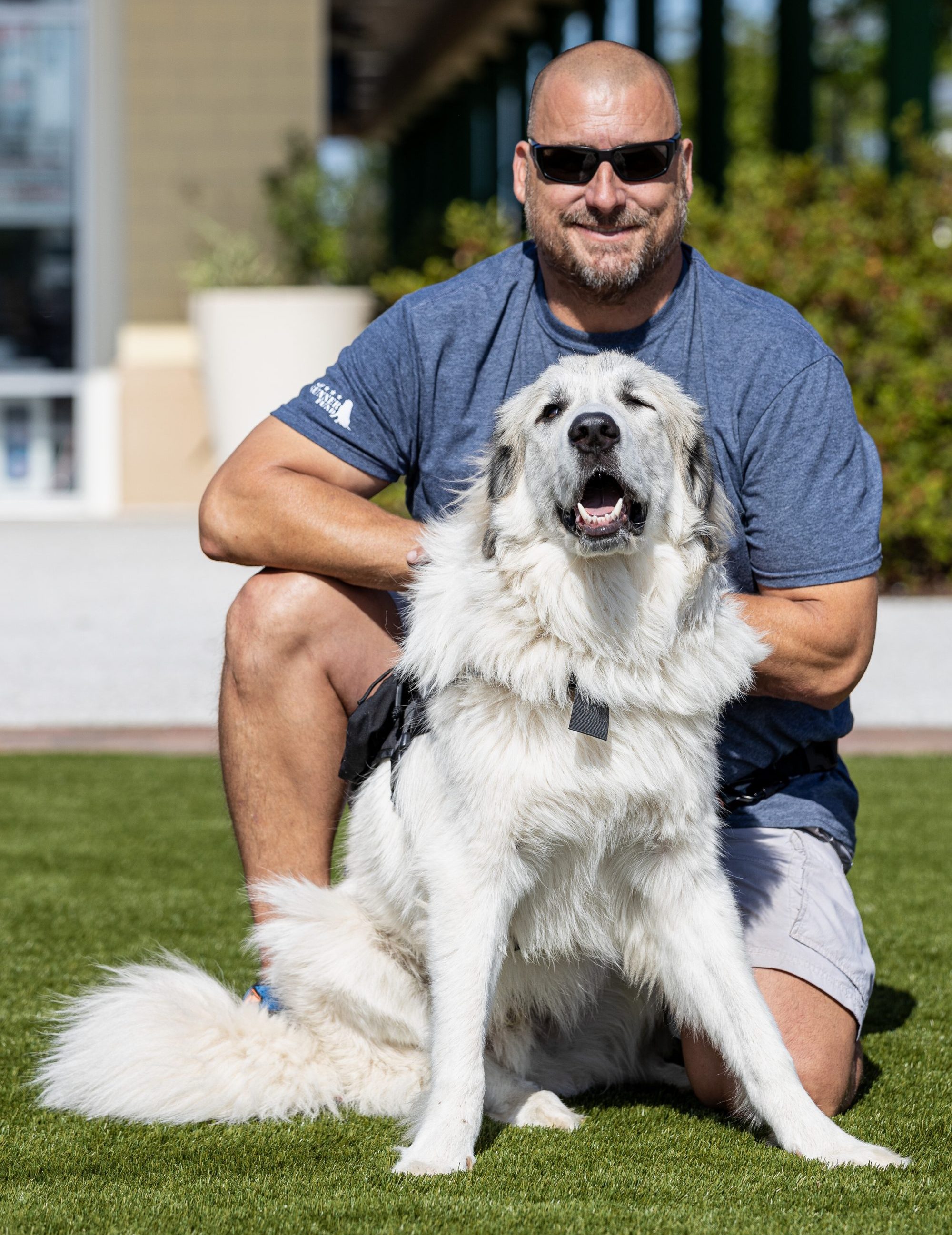Sergeant Hamilton Kinard, wearing dark sunglassess and a blue t shirt, sits behind his white Great Pyrenees service dog Gunner on the grass.