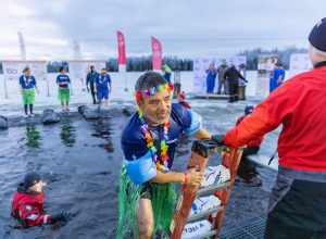 A wet Matson employee wearing floaties, a rainbow lei, and plastic grass skirt climbs a ladder to exit the chilly lake waters as lifeguards dressed in cold water gear keep watch.