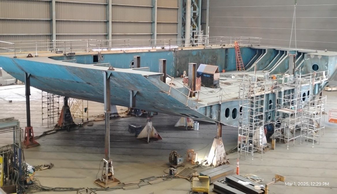 An unfinished section of the hull sits in a large shop braced by supports and surrounded by scaffolding.