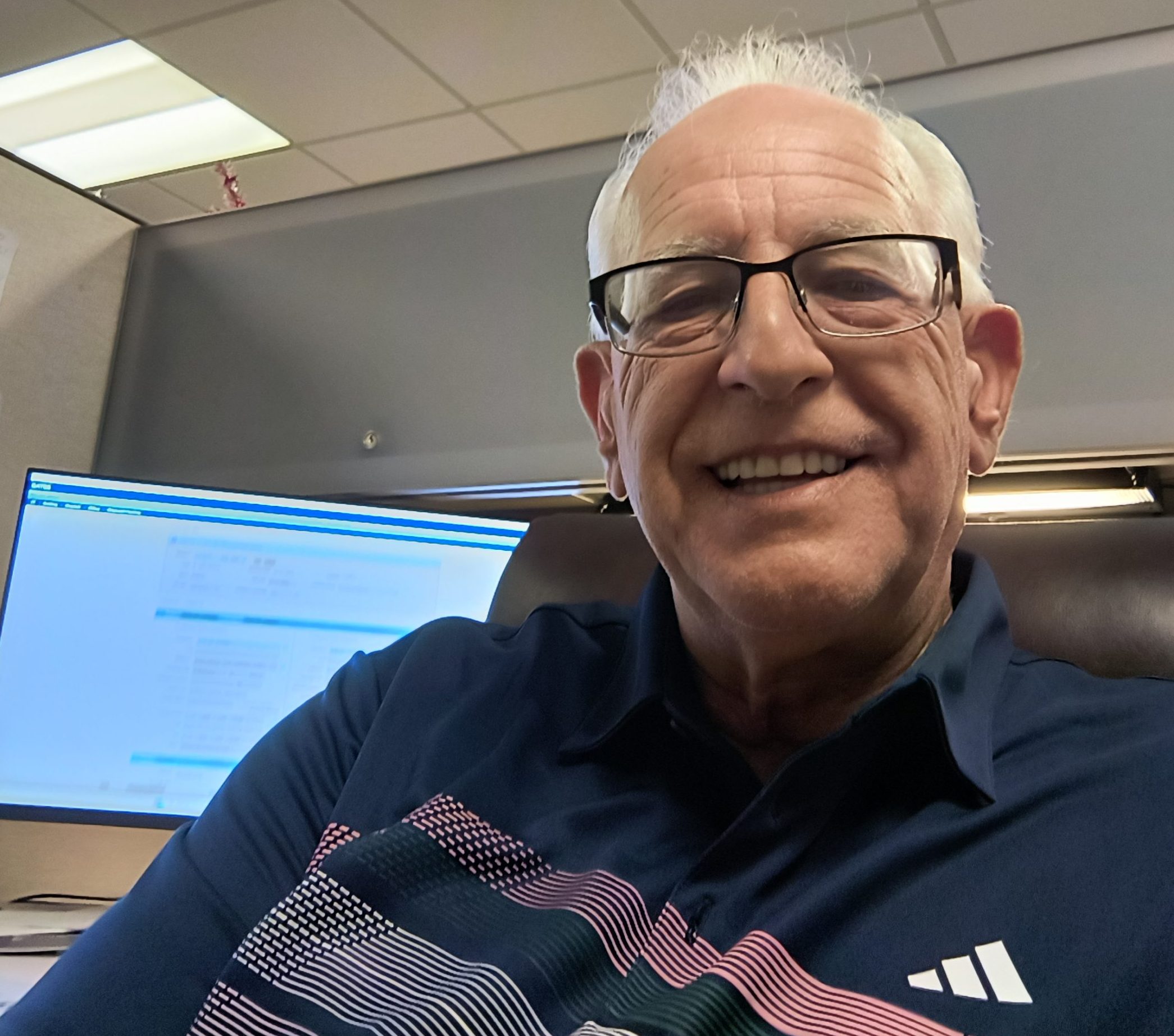Gabriel Basso sits in his Phoenix cubicle with a smile on his face. Overhead cabinets and a computer monitor are the background.