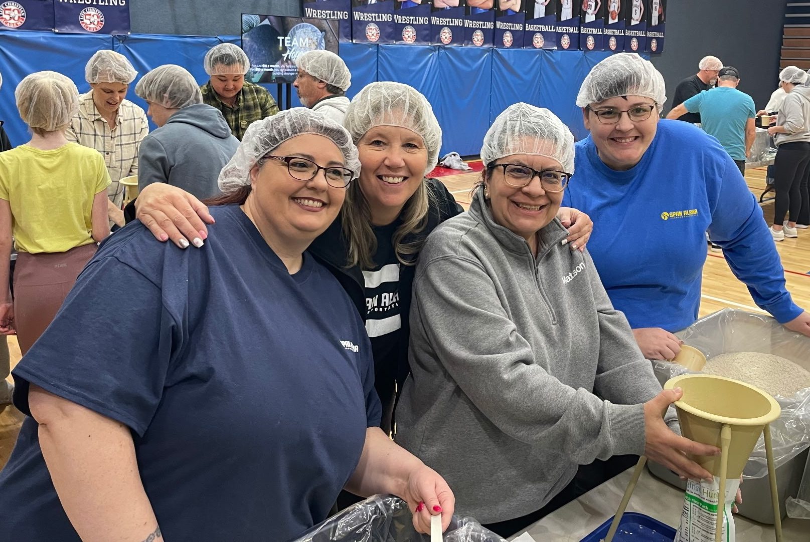 Span employees wearing white hair covers smile for the camera while they prepare food in a high school gym.